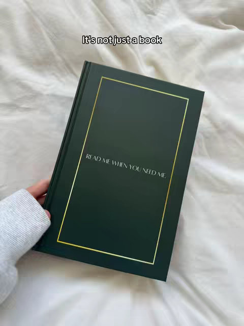 A hand holds a dark green book with a gold border. The book cover reads, 'READ ME WHEN YOU NEED ME.' The background is a soft, white, textured surface, likely a bed sheet.