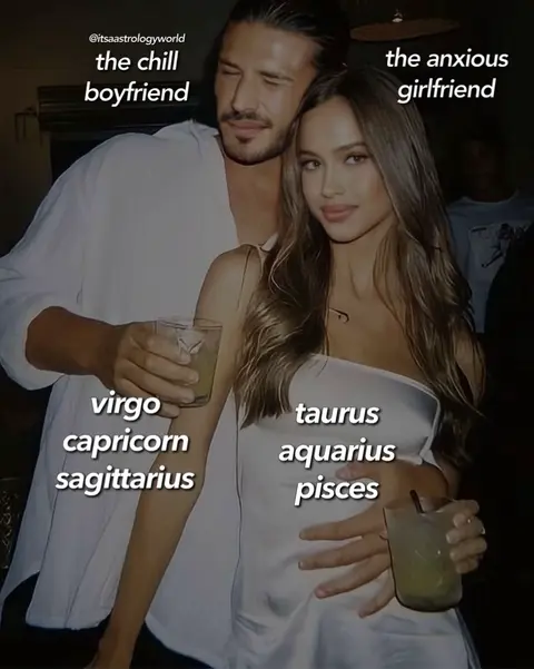 The image is a photograph of a couple standing together, likely in a social setting. The man is on the left, clad in a white, slightly oversized shirt. He has dark hair, and a shadow shows his side profile. The woman on the right has long, light brown hair and is wearing a white, strapless dress, holding a drink. Text overlays have been added to the image, identifying the couple as 'the chill boyfriend' and 'the anxious girlfriend', along with astrological signs associated with each. The background is dark, and the lighting is somewhat dim, highlighting the subjects.