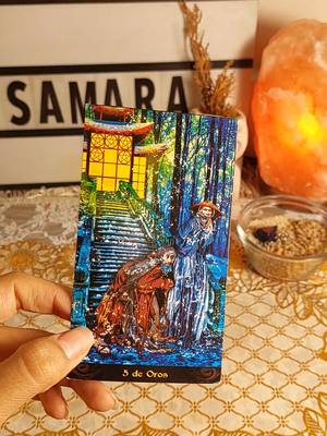 A hand holds a tarot card that depicts a scene with figures ascending and descending stairs, set against a backdrop of a forest. The card is the '5 de Oros'. The background features a lit sign with the name 'SAMARA' and a lit orange sphere.