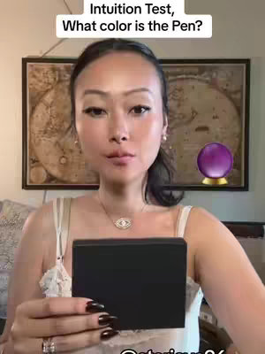A woman with long, dark hair and a white top is holding a black object in front of her chest. She has dark nail polish, eye makeup and is wearing an eye pendant. In the background are a painting, a crystal ball, and framed pictures.