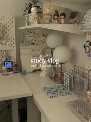 A well-lit, cozy desk setup with white furniture, shelves filled with decor, and a small digital clock. The scene is soft and inviting, with a study-themed atmosphere.