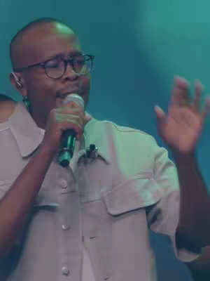 The image shows a man of color singing into a microphone. He is wearing glasses and has short hair. The background is a blurred blue-green.