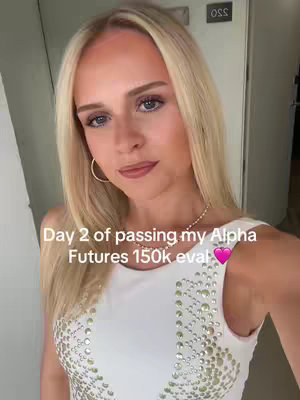 A selfie-style shot featuring a woman with long blonde hair, wearing a white top adorned with small, closely-spaced embellishments. The background includes a door and a wall. Text overlay includes "Day 2 of passing my Alpha Futures 150k eval", with a pink heart emoji.