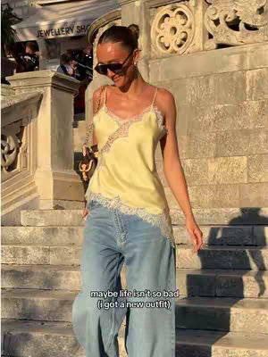 A woman in sunglasses and a yellow top walks down some stairs. She is wearing blue jeans and a small purse, posing for the photo in natural lighting. There is a sign for a jewelry shop in the background.