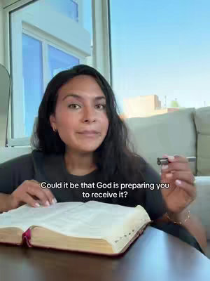 A woman with dark hair and a black shirt is seated at a wooden table with a Bible open in front of her. She's holding a pen and looking towards the camera with a gentle expression. The background features a window and a glimpse of an outdoor scene.