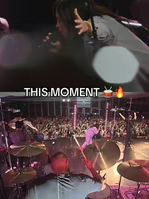 The cover image is a split view with two perspectives of a concert. In the upper portion, a person with long hair faces left, singing into a microphone with one arm raised. The bottom half shows a view of the stage with a drummer in the foreground and a large crowd of people in the background. The text "THIS MOMENT" with drum and fire emojis is superimposed.