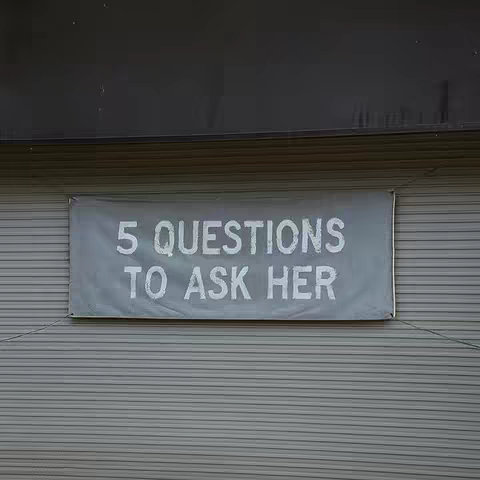 A banner with the text '5 QUESTIONS TO ASK HER' in white, sans-serif font hangs on a grey, horizontally-lined wall. The top of the image shows a darker, shadowed area.