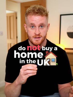 A man with fair skin and short, curly red hair is centered in the frame, wearing a black t-shirt. The shirt has white text overlaid across it, partially obscured by the man's pose, but still discernible. The background is blurred, suggesting a domestic interior with hints of wooden doors or cabinets and an off-white wall. The man's facial expression is neutral, looking directly at the camera.