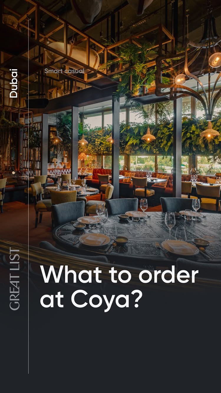 The cover image showcases a luxurious restaurant interior. A large, round, ornately designed table dominates the foreground, with multiple place settings featuring white plates, glassware, and small bowls. Behind the table, a series of smaller tables and booths are visible, enclosed by large glass windows that reveal a lush green outdoor setting. The restaurant's decor includes warm lighting, dark chairs and ceilings, and an emphasis on natural elements like plants. The text elements are well-integrated within the overall elegant composition.