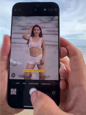 A hand holding a smartphone that is displaying a picture of a woman posing in a bikini at the beach. The woman is standing in shallow water, with her arms raised and the ocean and sky visible in the background.