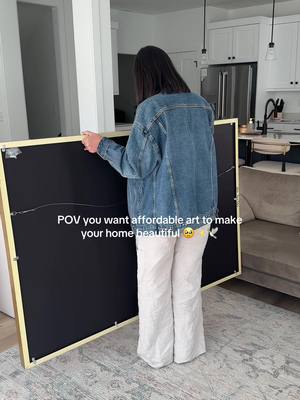 A woman, viewed from the back, holds a large, rectangular artwork in a modern home setting. She wears a denim jacket and light-colored pants. The artwork is unframed, revealing a wooden structure and a black canvas back. She's standing in front of a kitchen and living area, with a soft gray couch, white cabinetry, and a patterned rug