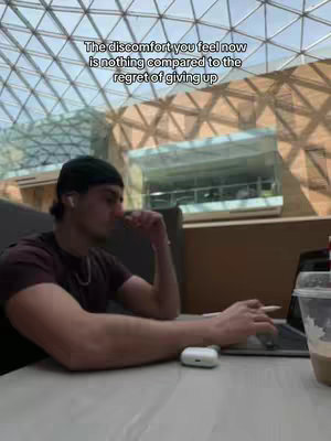 A man is shown sitting at a table, presumably in a public space with a high, glass-paneled ceiling. He is wearing a dark cap and a maroon shirt, with earbuds in his ears. He leans his arm on the table. In front of him is a laptop and a cup. Above the man, text declares, "The discomfort you feel now is nothing compared to the regret of giving up."