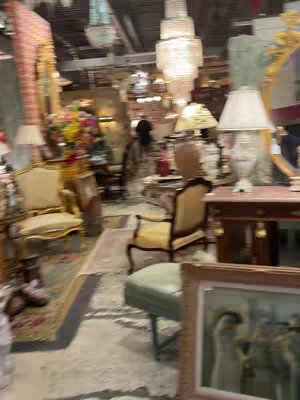 An interior shot of a shop filled with furniture and decorative items. A large chandelier hangs from the ceiling, and various lamps and furnishings are scattered throughout. A person is visible in the background.