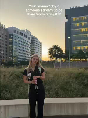 A woman in scrubs stands in front of a cityscape at dusk, holding a drink. Text overlay reads "Your \"normal\" day is someone's dream, so be thankful everyday".
