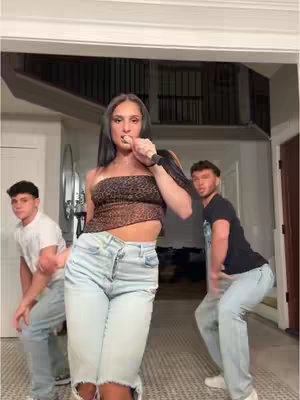A woman is in the center, holding a microphone and looking at the camera. Two men are dancing in the background. The room is well-lit with a light-colored interior.