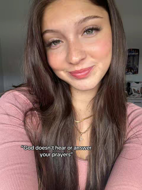 A selfie of a young woman with long brown hair, light makeup including mascara, and a simple gold cross necklace. She's smiling with pink lipstick, and a quote is overlaid on the image; the background is blurred.