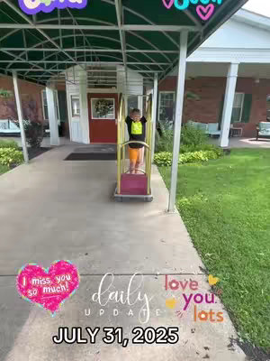 A young boy stands inside a yellow, metal-framed cart on a concrete pathway. The cart has what looks like a shelf at the bottom. The scene takes place in a brick building’s portico. Text overlay is present, featuring phrases like “I miss you so much!”, “Daily Update”, and the date.
