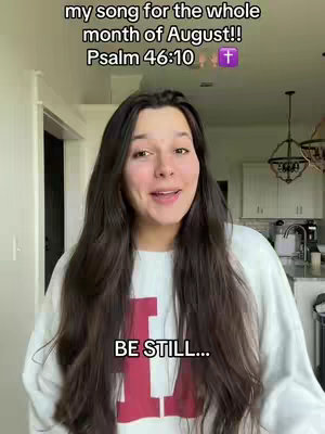 A woman with long, dark hair is centered in the frame, speaking. Above her, text overlays read: 'my song for the whole month of August!!' and 'Psalm 46:10' with a purple cross icon. Below her, the words 'BE STILL...' are displayed. The background shows a bright interior with a glimpse of a kitchen.