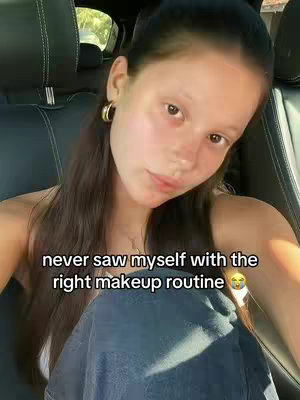 A young woman is seated in the driver's seat of a car, looking towards the camera. She has long, dark hair pulled back from her face, gold hoop earrings, and is wearing a white top. The text overlay reads, "never saw myself with the right makeup routine". The background shows parts of the car interior.