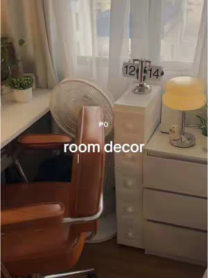 The image displays a well-lit room interior with a desk, chair, and various decorative items. A window with sheer curtains fills the background, providing natural light. A wooden desk and a brown leather chair are in the foreground, with a fan positioned to the left. On the right side, there's a white cabinet with a digital clock on top and a lamp. The color palette is soft and neutral, centered mostly on white and brown.