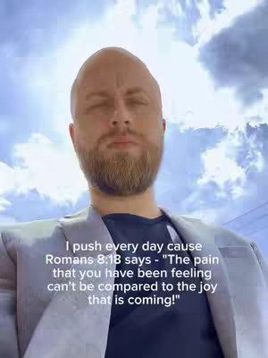 A man with a bald head and a beard is centered in the frame, wearing a blazer. The background shows a bright sky with sunlight.