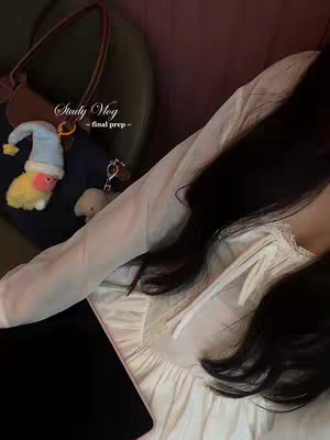 The image showcases a woman with long, dark hair, partially obscuring her face. She is wearing a white top and is angled in a relaxed, leaning position.  The background is blurred, suggesting an indoor setting, with a glimpse of a green surface and a bag with a cartoon keychains.