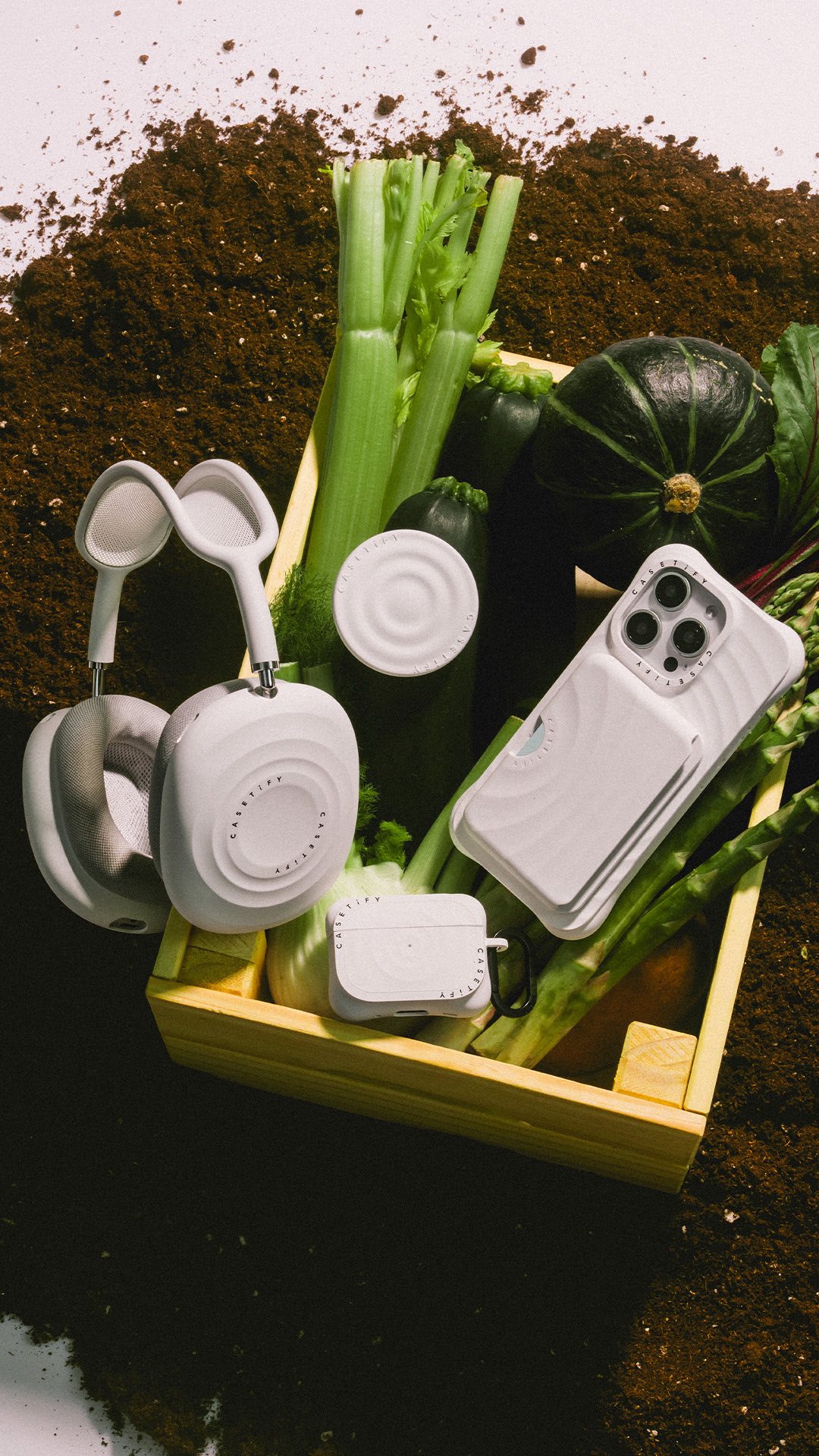 An overhead shot of several white Casetify accessories, including headphones and a wallet, arranged within a yellow wooden crate. The crate is filled with various green vegetables and leafy greens, set against a backdrop of dark soil.