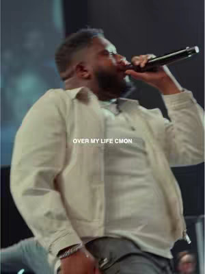 A man holding a microphone on a stage, singing. The image is lit with stage lights, and the background appears to be a dark stage. Overlayed text reads 'OVER MY LIFE CMON'.