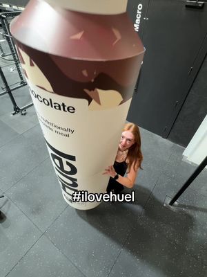 A young woman is standing behind a giant, oversized Huel bottle, posing and looking at the camera. The bottle dominates most of the frame, and the woman is partially hidden behind it, giving a playful scale comparison. The background suggests an indoor gym environment.
