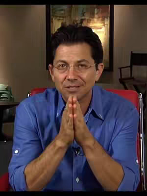 A middle-aged man with glasses sits in front of a dark background, a painting hangs behind him, and he wears a blue button-down shirt. He has dark hair. His hands are clasped together in front of his chest, seemingly in prayer or contemplation.