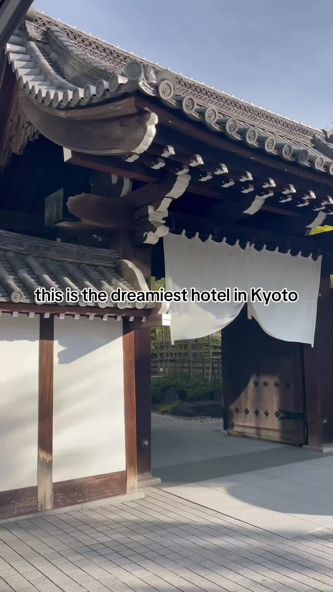 The image shows the entrance to a traditional Japanese building, possibly a hotel in Kyoto. The entrance features a large wooden gate with a tiled roof. A white curtain hangs down in the middle, and the text 'this is the dreamiest hotel in Kyoto' appears in a black, sans-serif font over the image. The scene is captured in daylight, with the composition emphasizing the aesthetic of a luxurious destination.