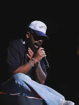A man wearing a white and navy baseball cap with text on it is holding a microphone and looking downward while sitting. He is wearing a dark t-shirt, light blue jeans, and a bracelet. The background is a dark, underexposed room.