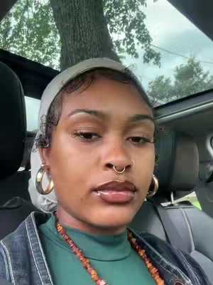 A selfie taken inside a car, featuring a woman with a headscarf and various accessories. The background includes a tree and the car's interior.