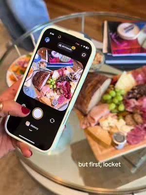 A person holding a phone that displays a photo of a charcuterie board. The phone is in front of the board. A sign on the phone displays the word "History". The hand is holding the phone. To the right is another charcuterie board.