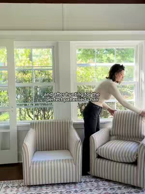 The image shows a sunroom with large windows and a woman interacting with a chair. The room has ample natural light, enhancing the airy feel. Two striped chairs are in the foreground, and the woman, dressed in a neutral-toned top and dark pants, is positioned near one of the chairs, possibly adjusting it. The neutral color palette of the room is complemented by the greenery visible through the windows.