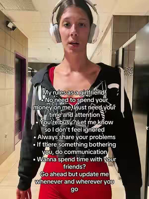 A woman stands in a tiled hallway, looking directly at the camera. She wears headphones, a red top, a black hoodie and red pants. Text overlays detail "My rules as a girlfriend".