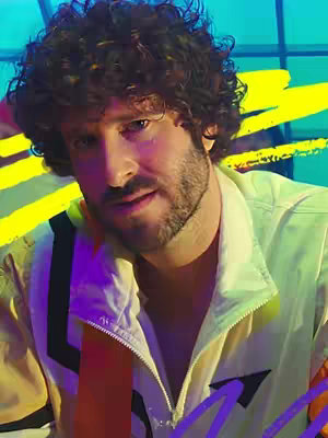 A man with curly brown hair and a beard is centered in a close-up shot, wearing a light yellow and black jacket. The background features a colorful, graffiti-style design with streaks of yellow, blue, and purple.