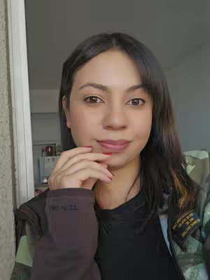 A woman with medium-length dark hair and warm skin tone poses for a selfie, looking directly at the camera with a slight smile. She's wearing a black top under a camouflage-patterned jacket. Her right hand rests near her face, with a few fingers touching her lower lip. The lighting is soft and natural, and the background is slightly blurred, suggesting an indoor setting, likely near a window.