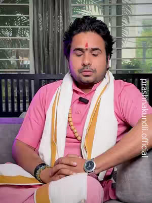 A man with a red dot on his forehead is sitting in a meditation pose with his eyes closed. He is wearing a pink shirt and a white shawl with gold accents. A wooden bead necklace is visible. The background features a window with blinds and a dark couch.