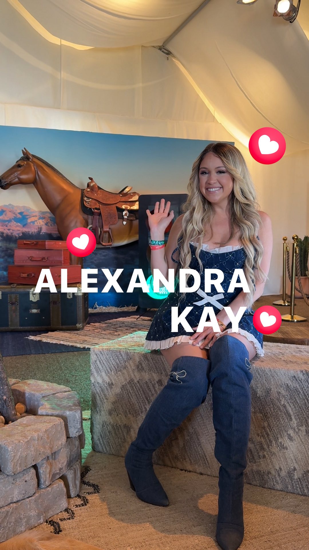 A promotional image featuring Alexandra Kay, a blonde woman in a denim dress and knee-high boots, waving. She is in a tent-like setting with a backdrop of a horse and travel trunks. Floating heart emojis and text overlays enhance the image.