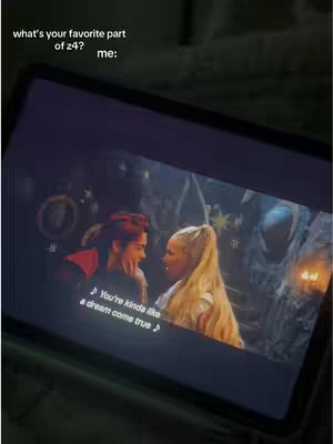 A screen of a device is visible, capturing a scene from the movie. Two characters are close to each other. One with red hair and  a black overcoat, other character with blonde hair and a dress. Both characters are looking at each other and smiling.