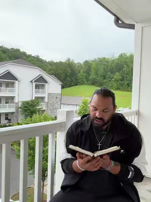 A man sits on a balcony, reading a book. Behind him is a view of trees, a grassy area, and a multi-story building. The man is holding the book in his lap. The balcony has white railings. The sky is overcast.