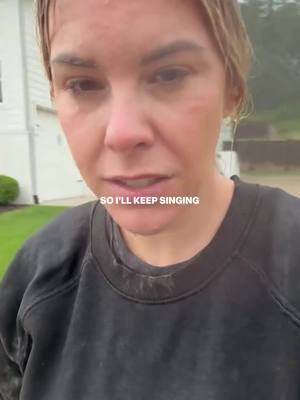 A woman stands outdoors in the rain, her face wet with raindrops. She's wearing a black sweatshirt and looking directly at the camera with a somewhat distressed expression. Overlay text reads "SO I'LL KEEP SINGING."