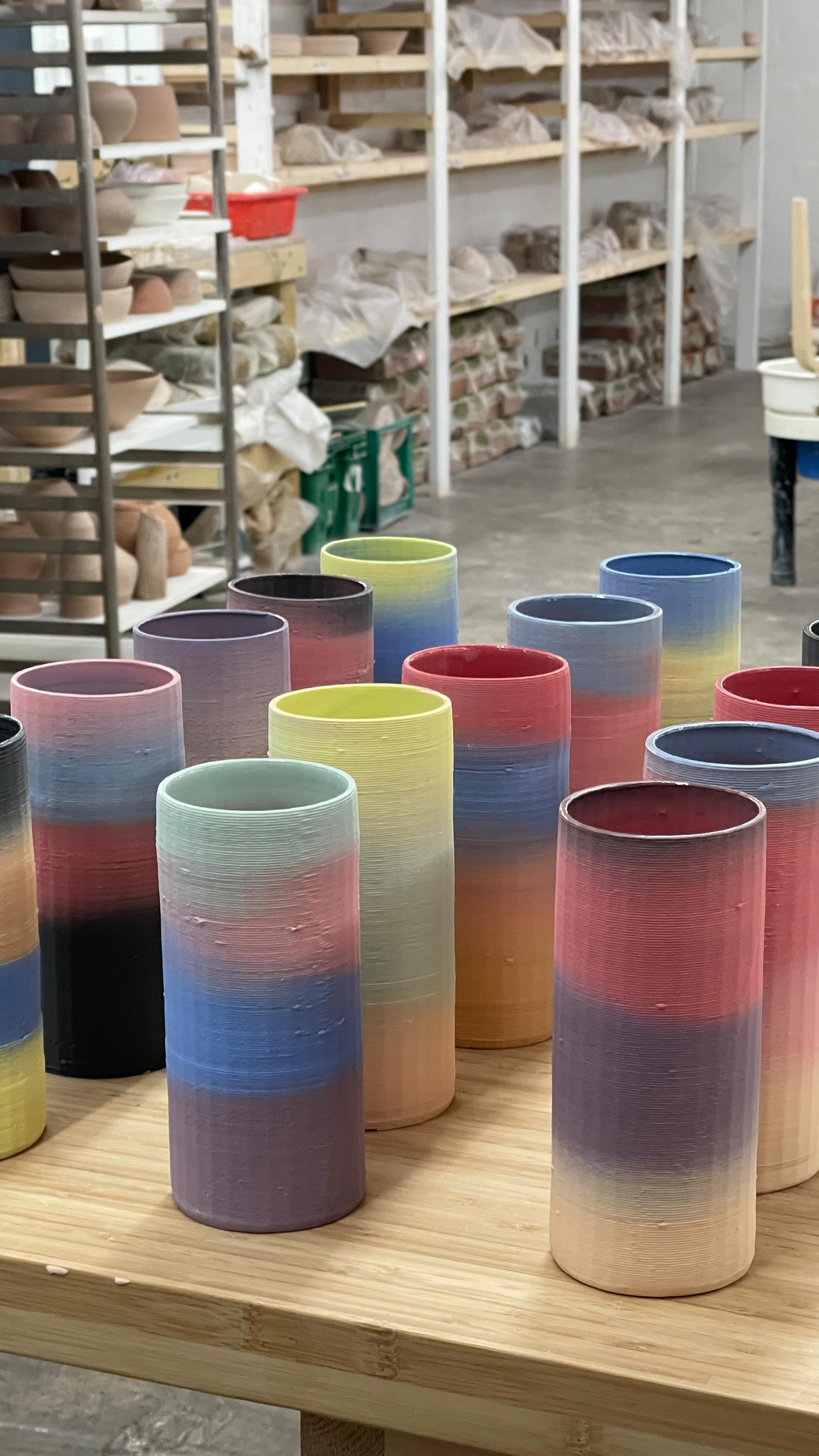 A close-up shot of several gradient-colored vases arranged on a wooden surface. The vases are cylindrical and display a smooth color transition from one shade to another. The background shows shelves filled with pottery items. The overall composition suggests a studio or workshop setting.