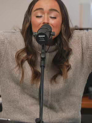 A woman is centered in the frame, singing into a microphone. She's wearing a light grey sweater with her arms slightly raised. Her eyes are closed, mouth open, and hair is styled with dark brown waves. A black microphone stand is in front of her.