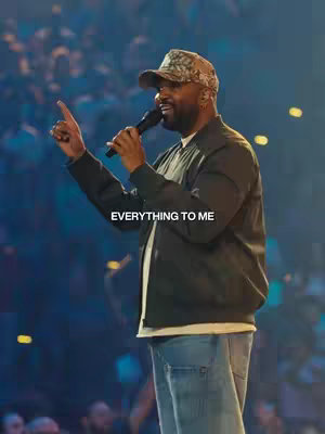 A medium shot featuring a Black man on stage, holding a microphone and singing. He's wearing a camo baseball cap, green jacket, and jeans; he's pointing to the sky with his left hand while holding a microphone in his right.