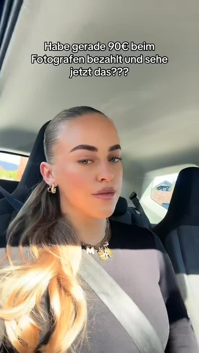A woman is seated in the driver's seat of a car, taking a selfie. She is wearing a black and grey top and a gold necklace. Her blonde hair is pulled back in a low ponytail. Text overlayed at the top says 'Habe gerade 90€ beim Fotografen bezahlt und sehe jetzt das???'.