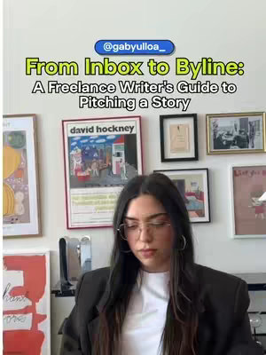A woman with long dark hair, wearing glasses and a blazer, is in front of a wall decorated with framed art. A sign reads From Inbox to Byline: A Freelance Writer's Guide to Pitching a Story.