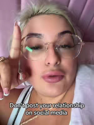 A close-up shot of a woman with blonde hair and glasses, pointing her index finger upwards. She has gold jewelry and is wearing a white top with a chain. Bottom of the frame reads "Don't post your relationship on social media."