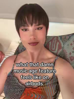 A woman with short, dark hair and expressive makeup appears in a selfie-style shot. She is wearing a patterned top, and a text overlay describes a feature on earbuds. The background is out of focus and suggests a domestic setting.